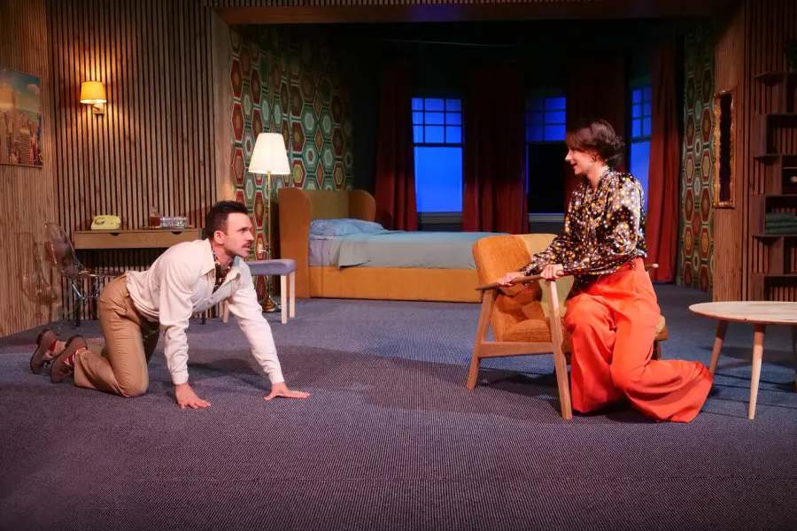 Na scenie teatralnej mężczyzna w koszuli i spodniach klęczy na podłodze, patrząc na siedzącą w fotelu kobietę w pomarańczowych spodniach i wzorzystej bluzce. W tle widoczna jest scenografia pokoju hotelowego z łóżkiem i drewnianymi panelami.