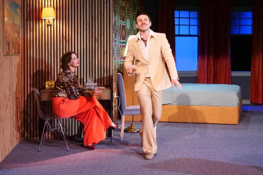 Na scenie teatralnej siedząca na krześle kobieta w pomarańczowych spodniach i wzorzystej bluzce trzyma szklankę, patrząc na stojącego obok mężczyznę w jasnym garniturze i z wąsem. Mężczyzna z uniesioną szklanką w ręku uśmiecha się szeroko, a w tle widać hotelowy wystrój z drewnianymi panelami.