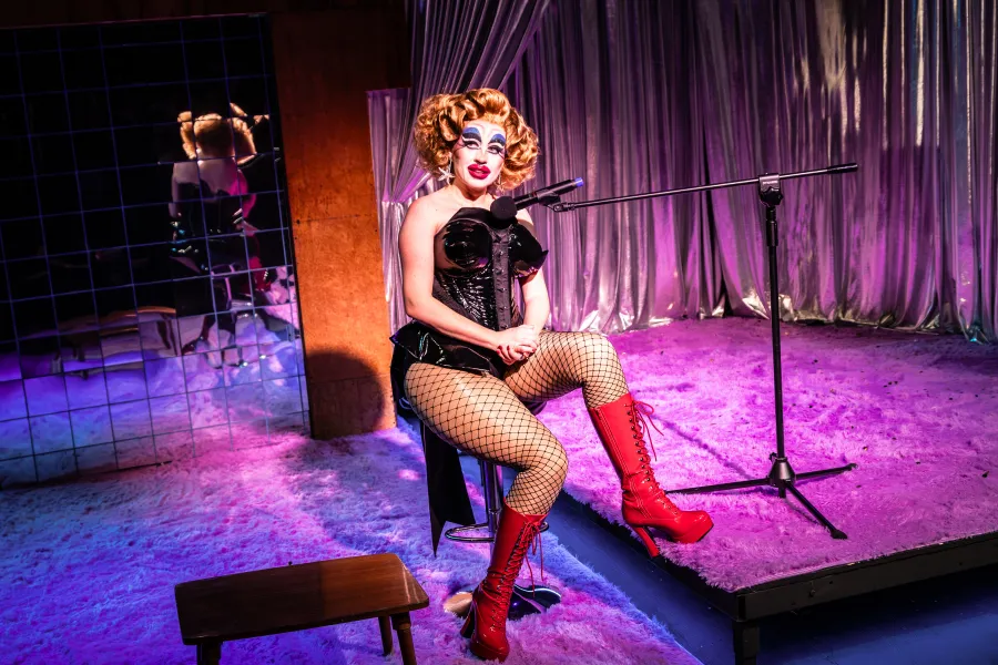 Zdjęcie ze spektaklu „Czas na wielki numer". Na fotografii widać aktorkę Matyldę Paszczenko ucharakteryzowaną na drag queen. Aktorka siedzi na stołku typu hoper. Do tworzy ma skierowany mikrofon. Ma na sobie czerwone, długie, skórzane kozaki na wysokim obsacie i lateksowy strój a la body. Aktorka ma wypchane uda i biodra. Ma na sobie także dużą, brązową perukę. Jej makijaż jest ostry, wyrazisty, wychodzący poza kontury twarzy. Autorką fotografii jest Natalia Kabanow