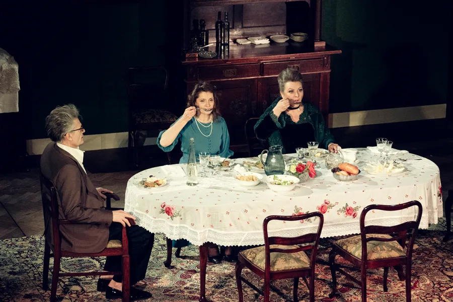 Na scenie teatralnej troje aktorów siedzi przy nakrytym stołem w jadalni, a dwie kobiety jedzą zupę. W tle widoczny jest ciemny kredens oraz obrazy na ścianie.