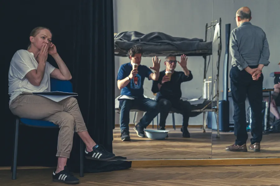 Krótkie nagranie lub próba teatralna w pomieszczeniu: na pierwszym planie siedzi kobieta z dokumentami, w tle dwaj mężczyźni gestykulują przy łóżku piętrowym, a tyłem stoi trzecia osoba. Scena oddaje atmosferę skupienia i pracy reżyserskiej/aktorskiej.