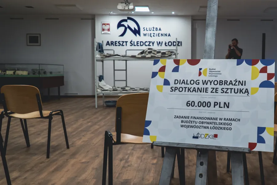 Na sztaludze na pierwszym planie stoi tablica informacyjna z napisem „DIALOG WYOBRAŹNI SPOTKANIE ZE SZTUKĄ, 60.000 PLN”. W tle widoczna jest sala z krzesłami, łóżko piętrowe oraz napis na ścianie „SŁUŻBA WIĘZIENNA ARESZT ŚLEDCZY W ŁODZI”.