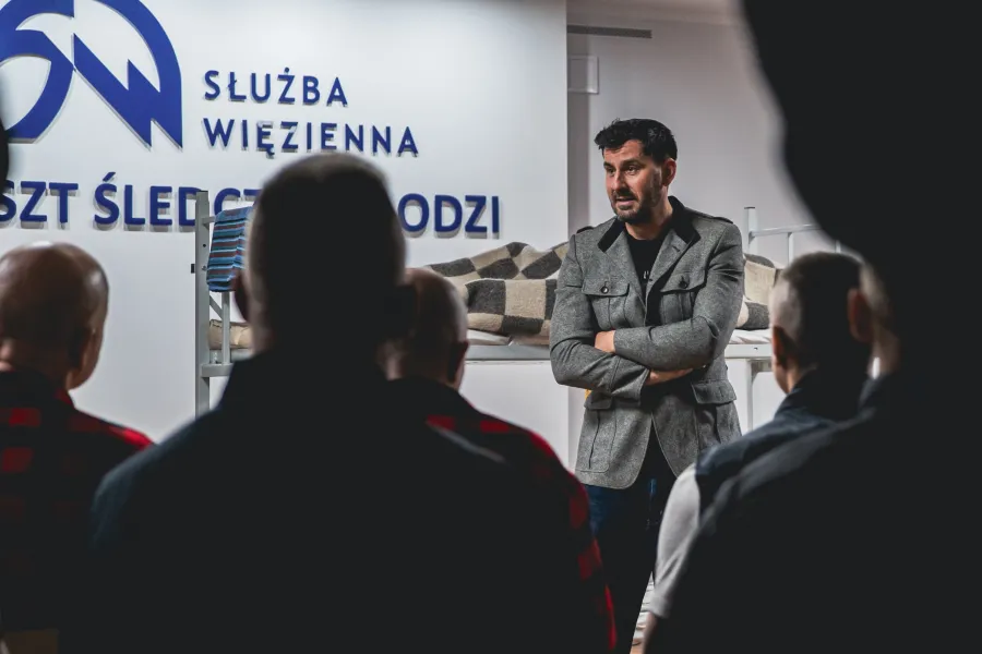 Mężczyzna w szarej kurtce z założonymi rękami stoi w pomieszczeniu przed grupą stojących tyłem osób. W tle widoczny jest napis „SŁUŻBA WIĘZIENNA” oraz fragment pryczy.