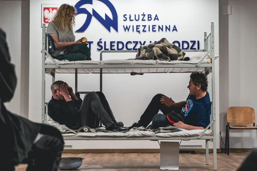 Trzy osoby siedzą na metalowym łóżku piętrowym w ramach prezentacji. W tle na ścianie widoczny jest baner z polskim godłem oraz napisami „SŁUŻBA WIĘZIENNA” i „ARESZT ŚLEDCZY W ŁODZI”.