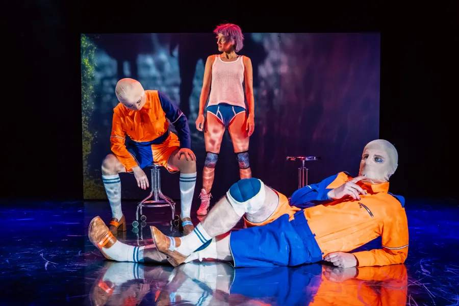 Trójka aktorów na ciemnej, oświetlonej scenie teatralnej. Po lewej siedzi osoba w pomarańczowo-niebieskim kombinezonie i z zasłoniętą twarzą. W centrum stoi kobieta w różowej peruce, podkoszulce i niebieskich majtkach. Z przodu po prawej leży na podłodze aktor w analogicznym kombinezonie, trzymający palec na ustach.