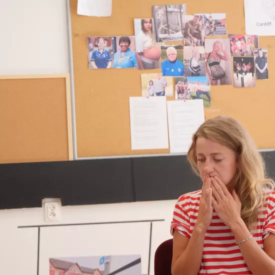 Zdjęcie przedstawia próbę stolikową do spektaklu „Ifigenia ze Splott”. Przy stole siedzi skupiona Agnieszka Skrzypczak z rękoma zasłaniającymi usta. Na stole oprócz zeszytu z ołówkiem znajduje się zdjęcie przedstawiające z tyłu brytyjską kamienicę, a z przodu znak z nazwą dzielnicy Splott. Z tyłu znajduje się korkowa tablica z wieloma zdjęciami różnych ludzi oraz napisem Cardiff.