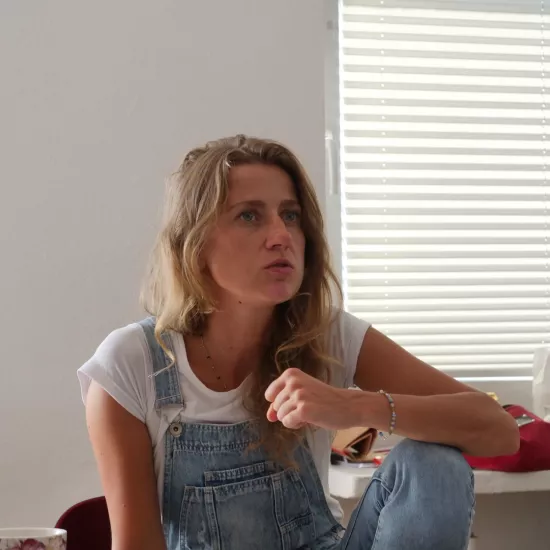 Zdjęcie przedstawia próbę stolikową do spektaklu „Ifigenia ze Splott”. Przy stole ze zgiętym kolanem, położonym na nim łokciu i zaciśniętą pięścią siedzi przy stole Agnieszka Skrzypczak. Aktorka patrzy przed siebie a kąciki jej ust są lekko opuszczone przy równoczesnym ich otwarciu. Przed aktorką na stole leży scenariusz, telefon, kubek w kwiaty, fotografie i długopis.  