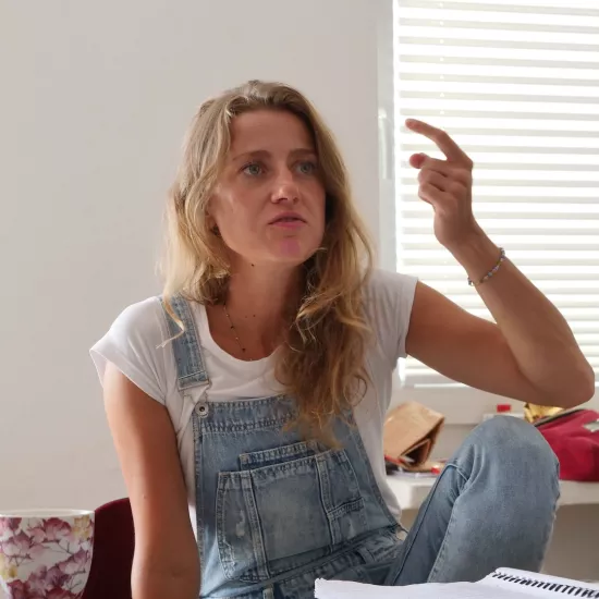 Zdjęcie przedstawia próbę stolikową do spektaklu „Ifigenia ze Splott”. Przy stole ze zgiętym kolanem i lekko podniesionym łokciem siedzi przy stole Agnieszka Skrzypczak. Wskazujący, zgięty palec aktorki jest lekko skierowany ku górze. Wzrok Agnieszki Skrzypczak skierowany jest przed siebie a kąciki jej ust są bardziej opuszczone w dół. Przed aktorką na stole z zielonym obrusem znajduje się scenariusz, fotografie, telefon, kubek w kwiaty i długopis.