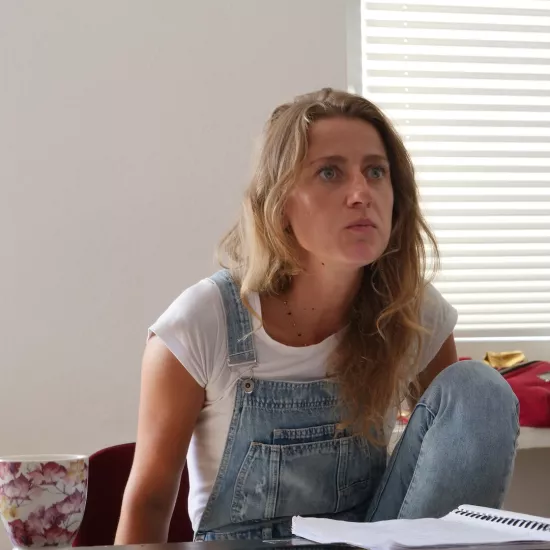 Zdjęcie przedstawia próbę stolikową do spektaklu „Ifigenia ze Splott”. Przy stole z lekko pochyloną do przodu, zgarbioną postawą i zgiętym kolanem siedzi Agnieszka Skrzypczak. Oczy aktorki są szeroko otwarte a kąciki jej ust lekko ściągnięte do środka. Na stole przed aktorką leży scenariusz, kubek w kwiaty, fotografie i telefon.