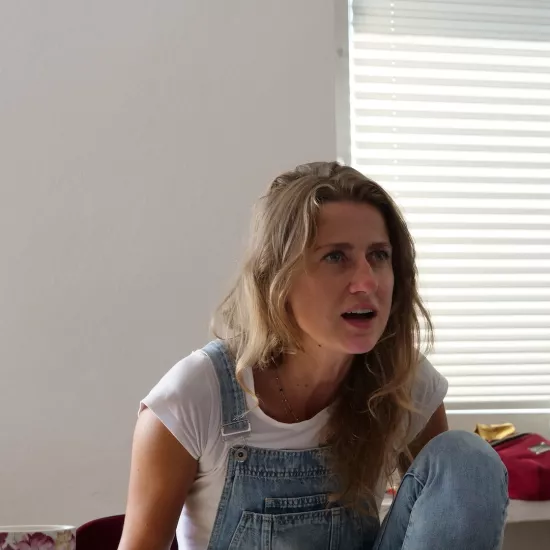 Zdjęcie przedstawia próbę stolikową do spektaklu „Ifigenia ze Splott”. Przy stole z lekko pochyloną do przodu, zgarbioną postawą i zgiętym kolanem siedzi Agnieszka Skrzypczak. Wzrok aktorki jest skierowany przed siebie, usta są otwarte a ręce zwisają wzdłuż ciała. Na stole przed aktorką leży scenariusz, kubek w kwiaty, fotografie i telefon.