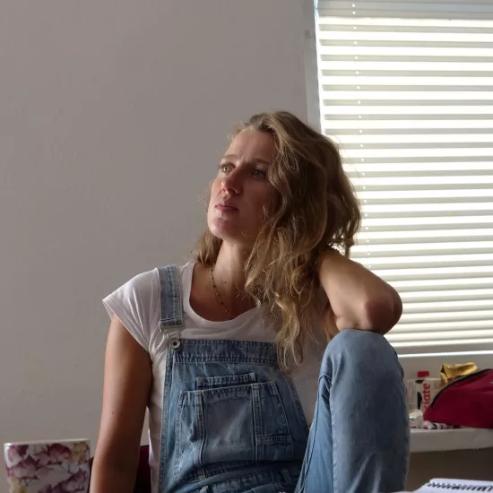 Zdjęcie przedstawia próbę stolikową do spektaklu „Ifigenia ze Splott”. Przy stole ze zgiętym kolanem i opartym na nim łokciu siedzi Agnieszka Skrzypczak. Ręka oparta na kolanie jest ugięta i sięga tyłu głowy aktorki a druga ręka zwisa wzdłuż ciała. Głowa Agnieszki Skrzypczyk jest zwrócona w prawą stronę i lekko pochylona do tyłu. W tę samą stronę, również sięga wzrok aktorki. Na stole przed aktorką leży scenariusz, kubek w kwiaty, fotografie i telefon.