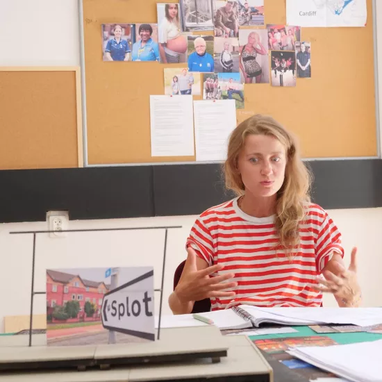  Zdjęcie przedstawia próbę stolikową do spektaklu „Ifigenia ze Splott”. Przy stole siedzi skupiona Agnieszka Skrzypczak z dwoma zgiętymi rękoma. Palce dłoni aktorki są mocno od siebie rozstawione a same dłonie wewnętrzną stroną do siebie lekko wyciągnięte do przodu. Oczy Agnieszki Skrzypczak zwrócone są poniżej linii obiektywu a kąciki jej ust mocno ściągnięte do siebie. Na stole oprócz zeszytu z ołówkiem znajduje się zdjęcie przedstawiające brytyjską kamienicę i znak z nazwą dzielnicy Splott.