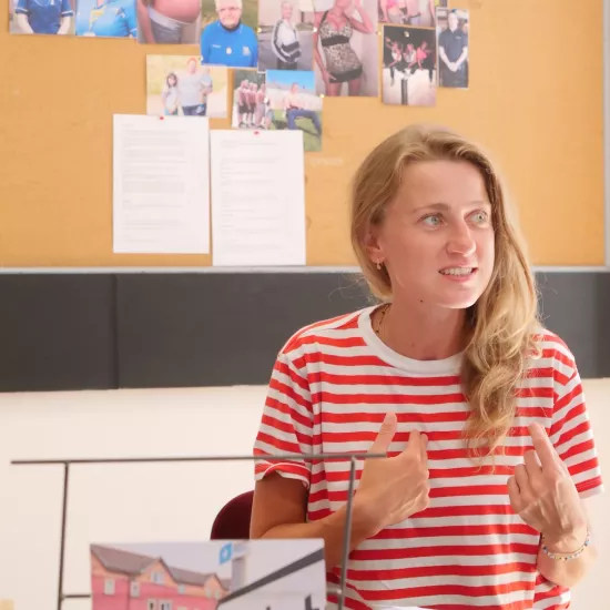 Zdjęcie przedstawia próbę stolikową do spektaklu „Ifigenia ze Splott”. Przy stole siedzi Agnieszka Skrzypczak ze zgiętymi w łokciach rękami skierowanymi na siebie. Wzrok aktorki jest skierowany poza obiektyw, a jej zęby są lekko zaciśnięte. Przed aktorką na stole znajdują się fotografie i makieta osiedlowego trzepaka. Na makiecie jest oparte zdjęcie angielskiej kamienicy z znakiem „Splott”. Oprócz trzepaka na stole znajduje się telefon, scenariusz i długopis. Z tyłu wisi na ścianie tablica korkowa.