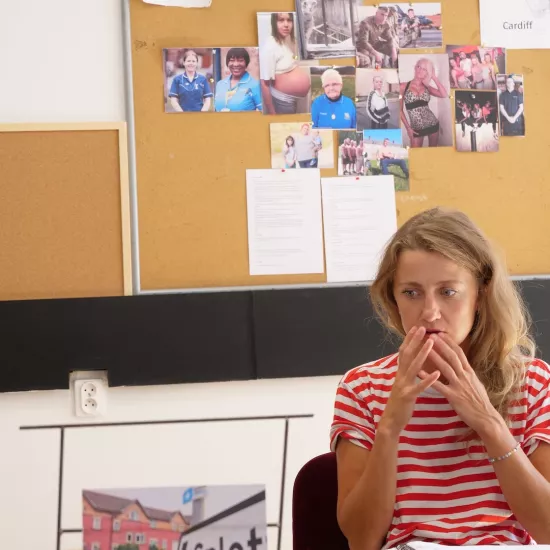 Zdjęcie przedstawia próbę stolikową do spektaklu „Ifigenia ze Splott”. Przy stole siedzi Agnieszka Skrzypczak ze złożonymi przy twarzy dłońmi. Wzrok aktorki jest skierowany poniżej obiektywu. Przed aktorką na stole znajdują się fotografie i makieta osiedlowego trzepaka. Na makiecie jest oparte zdjęcie angielskiej kamienicy z znakiem „Splott”. Oprócz trzepaka na stole znajduje się scenariusz i długopis. Z tyłu wisi na ścianie tablica korkowa ze zdjęciami.  