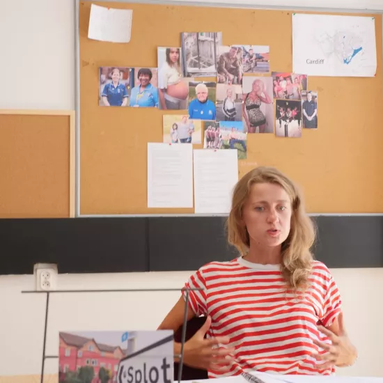 Zdjęcie przedstawia próbę stolikową do spektaklu „Ifigenia ze Splott”. Przy stole siedzi skupiona Agnieszka Skrzypczak z dwoma zgiętymi rękoma opartymi na oparciu krzesła. Palce dłoni aktorki są mocno od siebie rozstawione. Oczy Agnieszki Skrzypczak zwrócone są poniżej linii obiektywu a kąciki jej ust mocno ściągnięte do siebie. Na stole oprócz zeszytu z ołówkiem znajduje się zdjęcie przedstawiające z tyłu brytyjską kamienicę, a z przodu znak z nazwą dzielnicy Splott. 