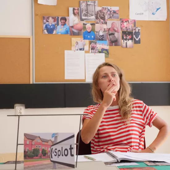 Zdjęcie przedstawia próbę stolikową do spektaklu „Ifigenia ze Splott”. Przy stole siedzi skupiona Agnieszka Skrzypczak z dwoma palcami na ustach i ze zgiętą ręką położoną na biodrze. Na stole oprócz zeszytu z ołówkiem znajduje się zdjęcie przedstawiające z tyłu brytyjską kamienicę, a z przodu znak z nazwą dzielnicy Splott. Z tyłu znajduje się korkowa tablica z wieloma zdjęciami różnych ludzi, tekstami oraz napisem Cardiff.