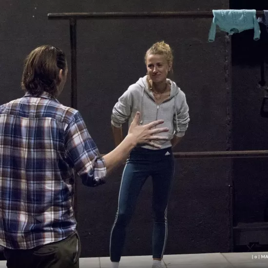 Zdjęcie przedstawia drugą próbę do spektaklu „Ifigenia ze Splott”. Agnieszka Skrzypczak stoi w centrum fotografii. Głowę ma lekką przechyloną w prawą stronę. Patrzy na Filipa Gieldona, który stoi plecami do kamery, po lewej stronie zdjęcia. Reżyser rozmawia z aktorką, przy czym gestykuluje rękoma. Aktorka ma ręce za plecami, lekko opiera się o scenografię w postaci trzepaka osiedlowego. Na górnej poprzeczce trzepaka, w prawym rogu, widać trzy sztuki błękitnej odzieży.