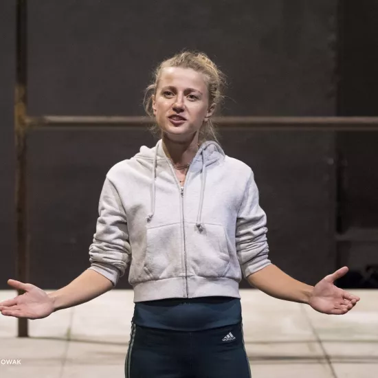 Zdjęcie przedstawia drugą próbę do spektaklu „Ifigenia ze Splott”. Agnieszka Skrzypczak stoi w centrum fotografii. Fotografia jest przybliżona, aktorkę widać od pasa w górę. Stoi na wprost obiektywu. Głowę ma skierowaną minimalnie w prawo, w tą stronę też patrzy. Ręce ma podniesione na wysokość bioder, dłonie otwarte, skierowane na zewnątrz. Aktorka ma otwarte usta. Za aktorką widać fragment lewej strony scenografii trzepaka osiedlowego na podeście.