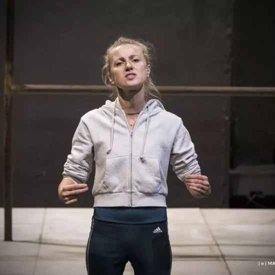 Zdjęcie przedstawia drugą próbę do spektaklu „Ifigenia ze Splott”. Agnieszka Skrzypczak stoi w centrum fotografii. Fotografia jest przybliżona, aktorkę widać od pasa w górę. Stoi na wprost obiektywu. Ręce ma podniesione na wysokość bioder. Wskazuje na siebie palcem wskazującym i środkowym prawej ręki, oraz lewą dłonią. Głowę ma lekko podniesioną, patrzy się w obiektyw. Usta ma nieznacznie otworzone. Za aktorką widać fragment lewej strony scenografii trzepaka osiedlowego na podeście. 