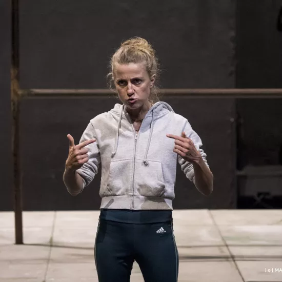 Zdjęcie przedstawia drugą próbę do spektaklu „Ifigenia ze Splott”. Agnieszka Skrzypczak stoi w centrum fotografii. Fotografia jest przybliżona, aktorkę widać od pasa w górę. Stoi na wprost obiektywu. Ręce ma nad biodrami. Wskazuje na siebie palcami wskazującymi obydwu rąk. Głowę ma skierowaną w prawo, w tą stronę też patrzy. Usta ma nieznacznie otworzone. Za aktorką widać fragment lewej strony scenografii trzepaka osiedlowego na podeście. 