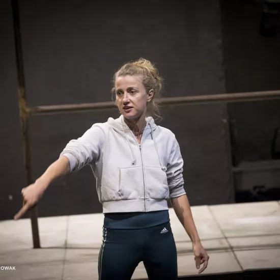 Zdjęcie przedstawia drugą próbę do spektaklu „Ifigenia ze Splott”. Agnieszka Skrzypczak stoi w centrum fotografii. Fotografia jest przybliżona, aktorkę widać od pasa w górę. Biodro aktorki jest lekko skręcone do boku. Aktorka ma głowę odwróconą w prawą stronę. Łokieć prawej ręki ma na wysokości klatki piersiowej, palcem wskazującym wskazuje lewy dolny róg fotografii. Wzrok aktorki jest skierowany w tą samą stronę co palec. Usta ma lekko otwarte. Za aktorką widać fragment lewej strony scenografii trzepaka.