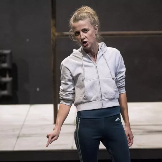 Zdjęcie przedstawia drugą próbę do spektaklu „Ifigenia ze Splott”. Agnieszka Skrzypczak stoi w centrum fotografii. Aktorka stoi na wprost obiektywu, lekko pochylona w prawą stronę. Głowę ma lekko odwróconą w prawo, wzrok ma skoncentrowany na obiektywie. Prawą rękę ma lekko zgiętą w łokciu, palcem wskazującym wskazuje na lewy dolny róg fotografii. Usta ma szeroko otwarte. Za aktorką widać fragment lewej strony scenografii trzepaka osiedlowego na podeście.