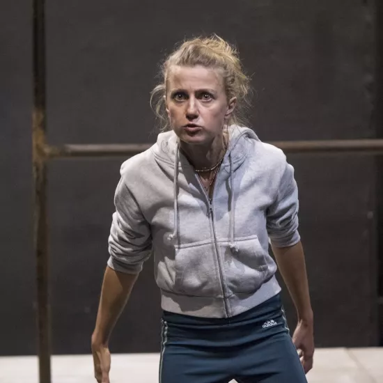 Zdjęcie przedstawia drugą próbę do spektaklu „Ifigenia ze Splott”. Agnieszka Skrzypczak stoi w centrum fotografii. Aktorka jest lekko pochylona w stronę obiektywu. Ręce ma nieznacznie za biodrami, dłonie ma otwarte. Nogi ma rozstawione. Aktorka patrzy się w obiektyw, oczy ma szeroko otwarte. Usta ma zamknięte, szczękę zaciśniętą. Za aktorką widać fragment lewej strony scenografii trzepaka osiedlowego na podeście.