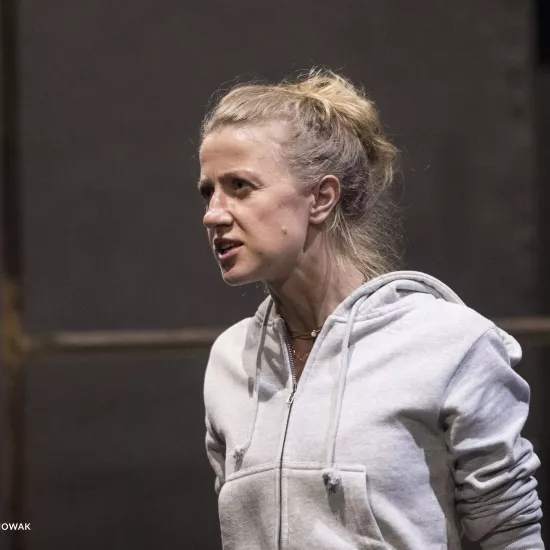 Zdjęcie przedstawia drugą próbę do spektaklu „Ifigenia ze Splott”. Agnieszka Skrzypczak stoi bokiem w centrum fotografii. Fotografia jest przybliżona, aktorkę widać od klatki piersiowej w górę. Aktorka patrzy w kierunku lewego końca fotografii. Usta ma otwarte, brwi ma zmarszczone. W tle widać fragment scenografii trzepaka osiedlowego.