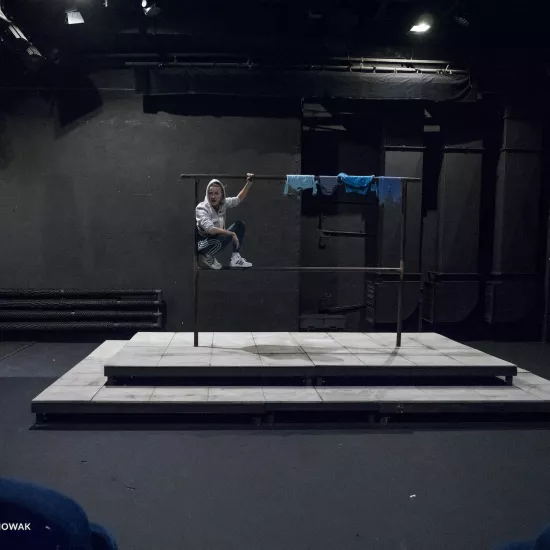 Zdjęcie przedstawia drugą próbę do spektaklu „Ifigenia ze Splott”. W centrum fotografii znajduje się scenografia trzepaka osiedlowego. Agnieszka Skrzypczak kuca na dolnej poprzeczce z lewej strony, opierając się plecami o lewy pręt. Lewą ręką trzyma górną poprzeczkę trzepaka, a prawą ma opartą na prawym kolanie. Jest lekko odwrócona w prawą stronę, nie patrzy w obiektyw. Na głowie ma założony kaptur białej bluzy. Na górnej poprzeczce trzepaka, z prawej strony, wiszą cztery sztuki błękitnej odzieży.