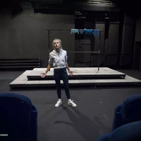 Zdjęcie przedstawia drugą próbę do spektaklu „Ifigenia ze Splott”. Agnieszka Skrzypczak stoi w centrum zdjęcia. Aktorka stoi na wprost do obiektywu, jest lekko pochylona do boku. Ręce ma lekko zgięte. Dłonie ma otwarte, skierowane wewnętrzną stroną do góry. Aktorka patrzy w obiektyw, głowę ma lekko podniesioną do góry. Ma otwarte usta. W dolnych rogach zdjęcia widać niebieskie fotele. Za aktorką widać scenografię trzepaka. Na górnej poprzeczce wisi błękitna odzież.