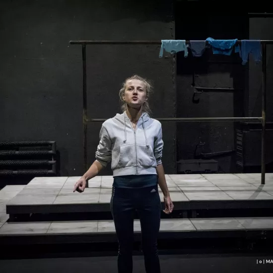 Zdjęcie przedstawia drugą próbę do spektaklu „Ifigenia ze Splott”. Agnieszka Skrzypczak stoi w centrum zdjęcia, przodem do obiektywu. Prawą rękę ma zgiętą, wskazuje palcem wskazującym w lewy dolny róg zdjęcia. Głowę ma lekko podniesioną do góry. Wzrok ma powyżej linii obiektywu. Usta ma lekko otwarte. Za aktorką widać scenografię trzepaka osiedlowego na podeście. Na górnej poprzeczce, po prawej stronie fotografii, wisi błękitna odzież.