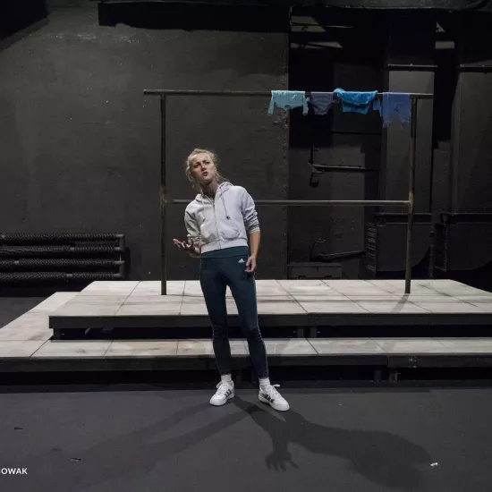 Zdjęcie przedstawia drugą próbę do spektaklu „Ifigenia ze Splott”. Agnieszka Skrzypczak stoi w centrum zdjęcia, przodem do obiektywu. Ciężar ciała aktorki skoncentrowany jest na prawej nodze. Aktorka jest lekko pochylona do tyłu. Prawą rękę ma przy ciele, zgiętą w łokciu. Prawą dłoń ma otwartą, wewnętrzną stroną do góry. Głowę ma podniesioną do góry. Wzrok ma powyżej linii obiektywu. Za aktorką widać scenografię trzepaka na podeście. Na górnej poprzeczce, po prawej stronie fotografii, wisi błękitna odzież.