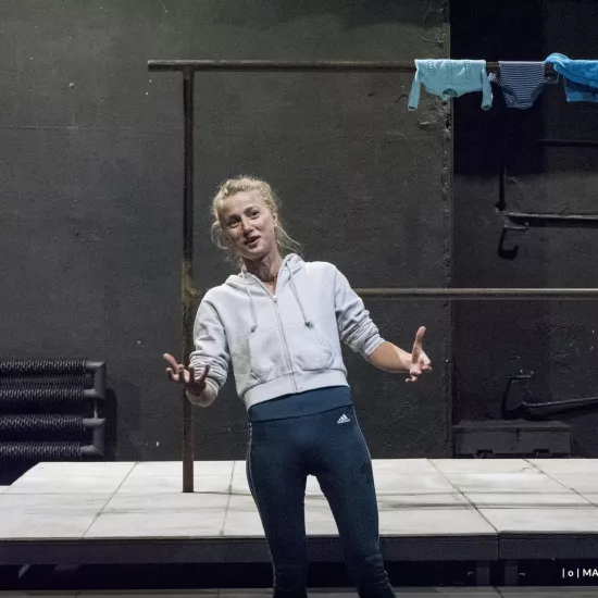 Zdjęcie przedstawia drugą próbę do spektaklu „Ifigenia ze Splott”. Agnieszka Skrzypczak stoi w centrum zdjęcia, przodem do obiektywu. Ciężar ciała aktorki skoncentrowany jest na prawej nodze. Aktorka jest lekko pochylona do tyłu. Ręce ma zgięte w łokciach. Dłonie ma otwarte, skierowane na zewnątrz. Wzrok ma skierowany w stronę prawego górnego rogu fotografii. Aktorka jest uśmiechnięta. Za aktorką widać scenografię trzepaka na podeście. Na górnej poprzeczce, po prawej stronie fotografii, wisi błękitna odzież