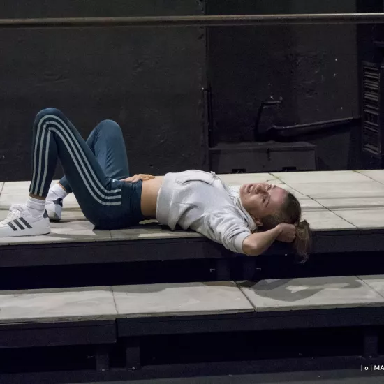 Zdjęcie przedstawia drugą próbę do spektaklu „Ifigenia ze Splott”. Agnieszka Skrzypczak znajduje się w centrum fotografii. Aktorka leży na podeście, lewym profilem do obiektywu. Aktorka ma głowę skierowaną w stronę obiektywu. Lewą ręką trzyma się za włosy, a prawą ma położoną na brzuchu. Nogi ma zgięte w kolanach. Aktorka jest uśmiechnięta. Za aktorką widać fragment scenografii osiedlowego trzepaka.