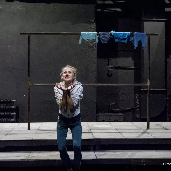 Zdjęcie przedstawia drugą próbę do spektaklu „Ifigenia ze Splott”. Agnieszka Skrzypczak znajduje się w centrum fotografii. Aktorka stoi na wprost obiektywu, jest pochylona do przodu, ma ugięte kolana. Ręce ma przed klatką piersiową, stykają się ze sobą. Dłonie są otwarte, skierowane wewnętrzną częścią w górę. Aktorka jest uśmiechnięta. Głowę ma podniesioną, wzrok skierowany ma w stronę lewego górnego rogu fotografii. Za aktorką widać scenografię trzepaka. Na górnej poprzeczce trzepaka wisi błękitna odzież.