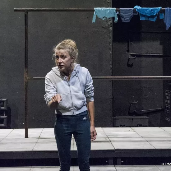 Zdjęcie przedstawia drugą próbę do spektaklu „Ifigenia ze Splott”. Agnieszka Skrzypczak znajduje się w centrum fotografii. Stoi na wprost obiektywu, jest lekko zgarbiona. Prawą dłoń ma podniesioną na wysokość bioder, palcem wskazującym wskazuje na obiektyw. Głowę ma odwróconą w prawo, aczkolwiek patrzy się w obiektyw. Usta ma lekko otworzone. Za aktorką widać scenografię trzepaka. Na górnej poprzeczce trzepaka, w prawym górnym rogu fotografii, wisi błękitna odzież.