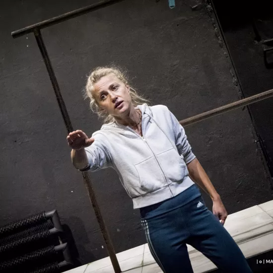 Zdjęcie przedstawia drugą próbę do spektaklu „Ifigenia ze Splott”. Agnieszka Skrzypczak znajduje się w centrum fotografii. Fotografia jest zrobiona pod kątem. Aktorka stoi na wprost obiektywu. Prawą dłoń ma na wysokości klatki piersiowej, wszystkimi palcami wskazuje na obiektyw. Patrzy w prawą stronę. Usta ma lekko otworzone. Za aktorką widać fragment scenografii osiedlowego trzepaka. 