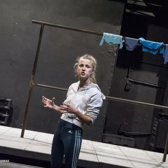 Zdjęcie przedstawia drugą próbę do spektaklu „Ifigenia ze Splott”. Agnieszka Skrzypczak znajduje się w centrum fotografii, stoi bokiem. Fotografia jest zrobiona pod kątem. Aktorka patrzy w obiektyw. Ręce ma zgięte pod kątem 90 stopni. Dłonie ma otwarte, skierowane wewnętrzną stroną do siebie. Usta ma lekko otwarte. Za aktorką widać scenografię trzepaka. Na górnej poprzeczce trzepaka wisi błękitna odzież.