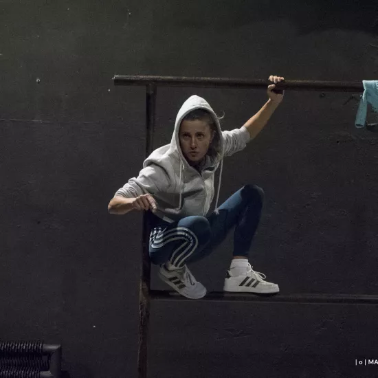 Zdjęcie przedstawia drugą próbę do spektaklu „Ifigenia ze Splott”. Agnieszka Skrzypczak kuca na scenografii trzepaka osiedlowego z lewej strony, opierając się plecami o lewy pręt. Lewą ręką trzyma górną poprzeczkę trzepaka, a prawą wskazuje w stronę kamery. Jest lekko odwrócona w prawą stronę, nie patrzy w obiektyw. Na głowie ma założony kaptur białej bluzy. W prawym rogu fotografii, na górnej poprzeczce widać zawieszoną błękitną bluzkę.