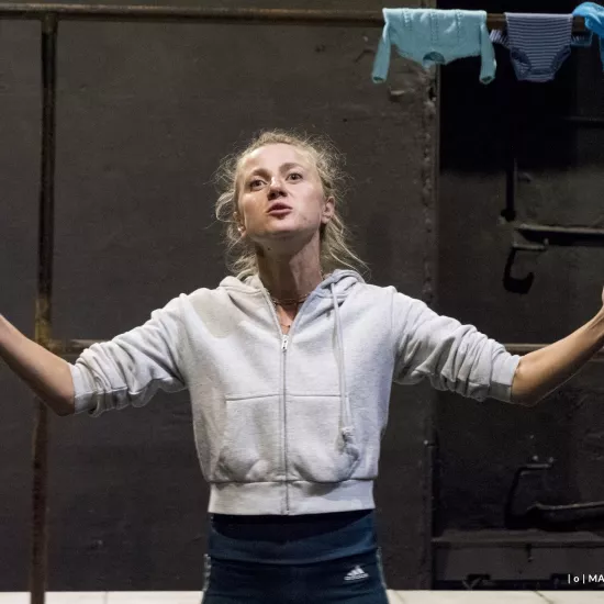 Zdjęcie przedstawia drugą próbę do spektaklu „Ifigenia ze Splott”. Agnieszka Skrzypczak znajduje się w centrum fotografii. Fotografia jest przybliżona, aktorkę widać od pasa w górę. Aktorka ma podniesioną głowę, patrzy ponad linię obiektywu. Ręce ma podniesione, szeroko otwarte, zgięte w łokciach. Dłonie ma otwarte wewnętrzną stroną w górę. Usta ma lekko otwarte. Za aktorką widać fragment scenografii trzepaka. Na górnej poprzeczce trzepaka wisi błękitna odzież.