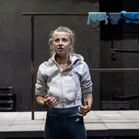 Zdjęcie przedstawia drugą próbę do spektaklu „Ifigenia ze Splott”. Agnieszka Skrzypczak znajduje się w centrum fotografii. Fotografia jest przybliżona, aktorkę widać od pasa w górę. Aktorka ma podniesioną głowę, patrzy ponad linię obiektywu. Ręce ma zgięte w łokciach. Palce ma lekko zgięte. Usta ma otwarte. Za aktorką widać fragment scenografii trzepaka. Na górnej poprzeczce trzepaka wisi błękitna odzież.