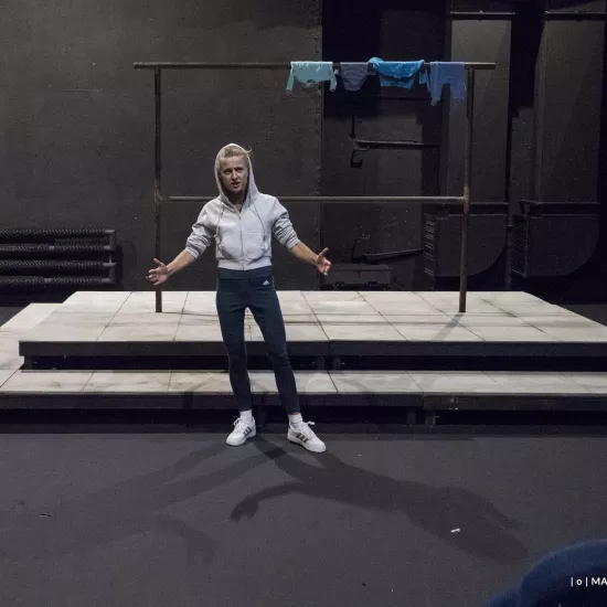 Zdjęcie przedstawia drugą próbę do spektaklu „Ifigenia ze Splott”. Przed podestem, na którym znajduje się scenografia trzepaka osiedlowego, stoi Agnieszka Skrzypczak. Ręce ma podniesione na wysokość bioder, rozłożone na boki. Jest lekko pochylona w prawo, lewą nogę ma lekko wysuniętą w bok. Patrzy się w obiektyw. Ma lekko otwarte usta. Na głowie ma założony kaptur białej bluzy. Na górnej poprzeczce trzepaka znajdującego się za nią, z prawej strony zawieszone są cztery sztuki błękitnej odzieży.