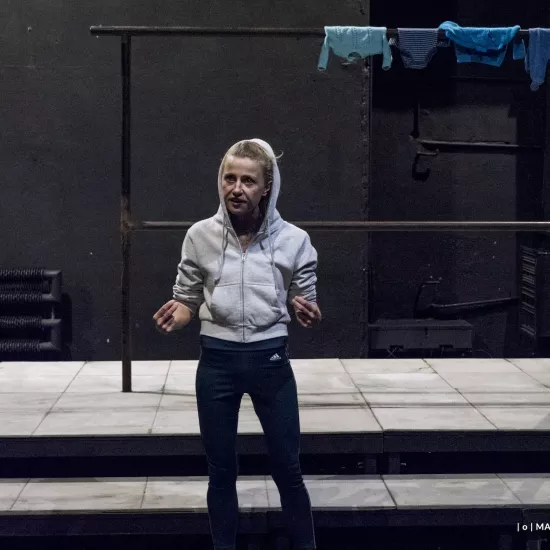 Zdjęcie przedstawia drugą próbę do spektaklu „Ifigenia ze Splott”. Przed podestem, na którym znajduje się scenografia trzepaka osiedlowego, stoi Agnieszka Skrzypczak. Ręce ma podniesione na wysokość bioder. Wskazuje na siebie palcami wskazującymi i środkowymi obydwu rąk. Patrzy w prawą stronę, nie w obiektyw. Usta ma lekko otwarte. Na głowie ma założony kaptur białej bluzy. Na górnej poprzeczce trzepaka znajdującego się za nią, z prawej strony zawieszone są cztery sztuki błękitnej odzieży. 