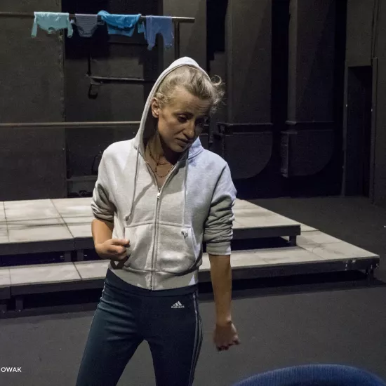 Zdjęcie przedstawia drugą próbę do spektaklu „Ifigenia ze Splott”. Agnieszka Skrzypczak stoi w centrum fotografii. Głowę ma lekko odwróconą w lewo, wzrok ma skierowany na podłogę. Prawą ręką trzyma kieszeń bluzy. Prawą nogę ma wysuniętą lekko w bok. Na głowie ma założony kaptur białej bluzy. Z lewej strony fotografii, za nią, stoi scenografia osiedlowego trzepaka na podeście. Na górnej poprzeczce trzepaka, z prawej strony zawieszone są cztery sztuki błękitnej odzieży.