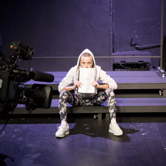 Zdjęcie przedstawia pierwszą próbę do spektaklu „Ifigenia ze Splott”. Na podeście w rozkroku siedzi Agnieszka Skrzypczak. Pod nosem trzyma scenariusz, przodem do obiektywu. Aktorka ubrana jest w ciemne legginsy w kwiaty, białe buty sportowe i białą bluzę, której kaptur ma założony na głowę. Uszy Agnieszki Skrzypczak ozdobione są dużymi kolczykami w kształcie okręgów w kolorze złota a włosy mocno zaczesane do tyłu. Za aktorką znajduje się scenografia w postaci trzepaka osiedlowego a przed nią kamera.  