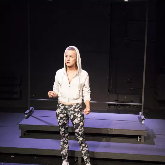 Zdjęcie przedstawia pierwszą próbę do spektaklu „Ifigenia ze Splott”. Przed podestami stoi Agnieszka Skrzypczak. Prawa ręka aktorki jest podniesiona na wysokość biodra a lewa na wysokość pępka. Wzrok Agnieszki Skrzypczak skierowany jest lekko do boku od obiektywu a jej usta delikatnie otwarte. Aktorka ubrana jest w ciemne legginsy w kwiaty, białe buty sportowe i białą bluzę, której kaptur ma założony na głowę. Za aktorką znajduje się scenografia w postaci trzepaka osiedlowego.