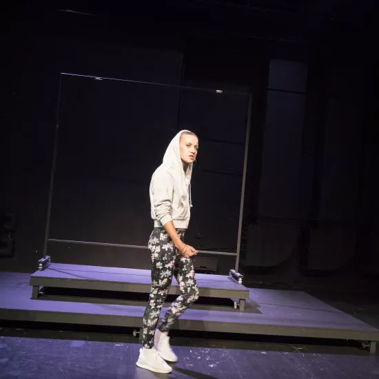 Zdjęcie przedstawia pierwszą próbę do spektaklu „Ifigenia ze Splott”. Przed podestami, bocznym profilem stoi Agnieszka Skrzypczak. Lewa noga aktorki jest zgięta w kolanie a prawa wyprostowana. Prawa ręka aktorki dotyka jej lewego uda a całą postawa jest lekko zgarbiona. Wzrok Agnieszki Skrzypczak sięga linii obiektywu, jej usta są otwarte a żuchwa lekko wysunięta do przodu. Aktorka ma założony kaptur na głowę a za nią znajduje się scenografia w postaci trzepaka osiedlowego.