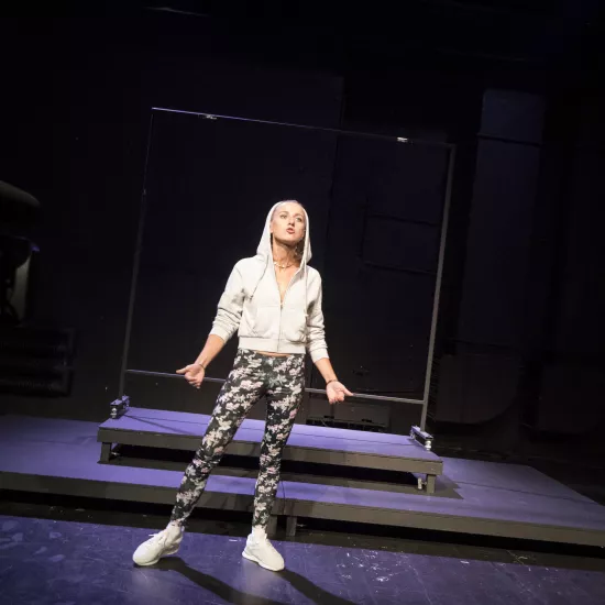 Zdjęcie przedstawia pierwszą próbę do spektaklu „Ifigenia ze Splott”. Przed podestami stoi Agnieszka Skrzypczak. Jej prawa noga jest odstawiona do boku i lekko zgięta w kolanie. Ręce aktorki są zgięte w łokciach i lekko podniesione wewnętrznymi częściami dłoni do góry. Oczy Agnieszki Skrzypczak są wytrzeszczone, głowa pochylona lekko do tyłu a usta mocno ściągnięte. Aktorka ma założony kaptur na głowę a za nią znajduje się scenografia w postaci trzepaka osiedlowego.