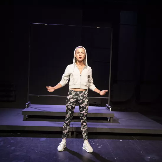 Zdjęcie przedstawia pierwszą próbę do spektaklu „Ifigenia ze Splott”. Przed podestami stoi Agnieszka Skrzypczak. Ciężar ciała aktorki jest rozstawiony na obu wyprostowanych nogach a jej miednica jest wysunięta do przodu. Ręce Agnieszki Skrzypczak są zgięte w łokciach i rozłożone. Wewnętrzne części dłoni są skierowane do góry. Wzrok aktorki jest w linii obiektywu a jej usta są lekko otwarte. Aktorka ma założony kaptur na głowę a za nią znajduje się scenografia w postaci trzepaka osiedlowego.