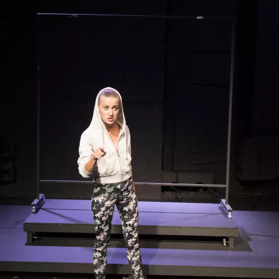 Zdjęcie przedstawia pierwszą próbę do spektaklu „Ifigenia ze Splott”. Przed podestami stoi Agnieszka Skrzypczak. Biodro aktorki jest lekko skręcone do boku, jej wzrok jest skierowany przed siebie poniżej linii obiektywu. Prawa ręka jest zgięta w łokciu a palec wskazujący tej ręki jest wyciągnięty przed siebie. Oczy Agnieszki Skrzypczak są mocno otwarte a jej usta otwarte z dużym uwzględnieniem dolnej wargi ust. Aktorka ma założony kaptur na głowę a za nią znajduje się scenografia w postaci trzepaka.